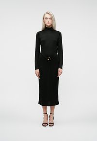 Blondína vo čiernych dlhých rukávoch svetrových �šatách s čiernym opaskom so zlatým akcentom a čiernymi sandálmi na vysokom opätku stojí na jednoduchom pozadí.