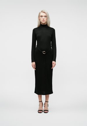 Mulher loira em um vestido preto de gola alta de mangas compridas, com um cinto preto com detalhes em dourado e sapatos de salto alto pretos com tiras, de pé sobre um fundo simples.