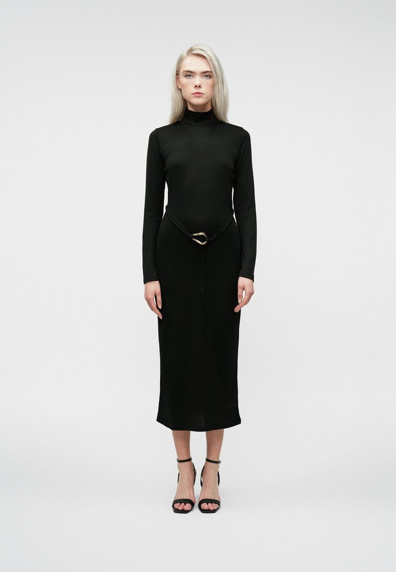 Blondína vo čiernych dlhých rukávoch svetrových šatách s čiernym opaskom so zlatým akcentom a čiernymi sandálmi na vysokom opätku stojí na jednoduchom pozadí.