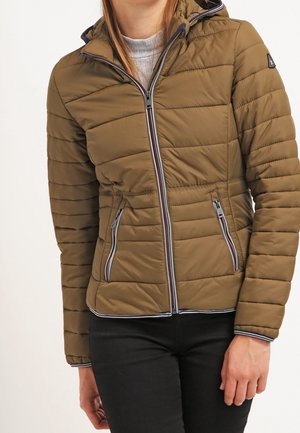 Femme portant une veste matelassée marron avec une fermeture éclair à l’avant et des poches zippées, associée à un pantalon noir.