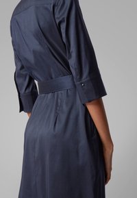 Granatowa sukienka wykonana z gładkiego materiału, z dopasowanym wcięciem w talii, rękawami o długości trzech ćwierć oraz paskiem, z subtelnymi fałdami z tyłu.
