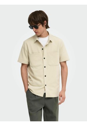 Mann trägt beige kurzärmliges Hemd mit Knöpfen, weißes Unterhemd, dunkle Hose und schwarze Sonnenbrille, steht mit den Händen in den Taschen und schaut nach unten.