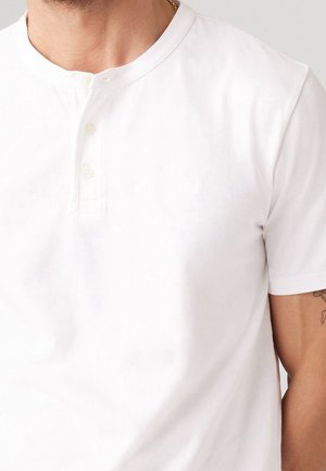 Homme portant un t-shirt henley blanc uni à manches courtes avec trois boutons, un tatouage visible sur le haut du bras et un collier en or autour du cou.