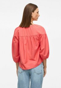 Koralfarvet skjorte lavet af glat stof, med en samlet ryg, bluseærmer og en løs pasform. Matchet med lyseblå jeans.