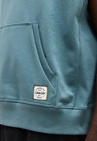 Primer plano del bolsillo de una sudadera con capucha Nike color verde azulado con una etiqueta blanca cosida que dice "Athletic Club, Established 1972, Just Do It." sobre un brazo de piel oscura.