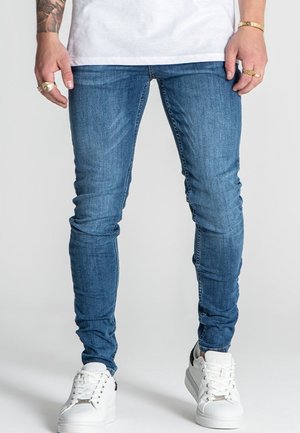 Uomo che indossa jeans blu aderenti, sneakers bianche, anelli d'oro, un braccialetto e una maglietta bianca, in piedi su uno sfondo semplice.