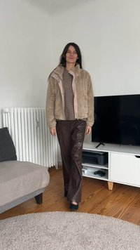 Beige faux fur jas met rits, bruine gebreide trui en bruine neplederen broek. Zwarte instapschoenen. Eenvoudig, neutraal kleurenpalet.
