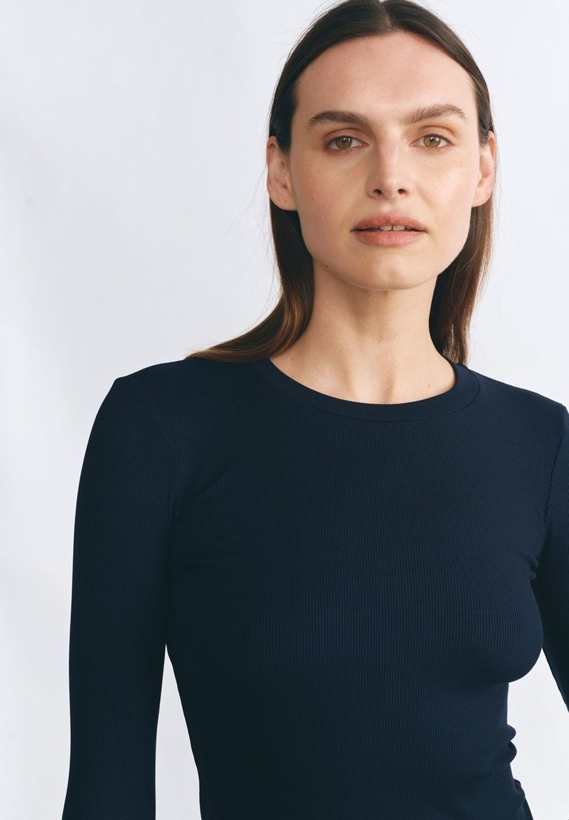 Kobieta z prostymi brązowymi włosami, ubrana w dopasowaną, długą, niebieską ribbed bluzkę na tle gładkiego jasnego tła.