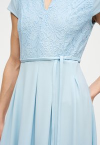 Φόρεμα ανοιχτού γαλάζιου χρώματος με μπούστο από δαντέλα με λουλούδια, κοντά μανίκια, εφαρμοστή μέση με λεπτή ζώνη και πλισέ φούστα κάτω από τη μέση.
