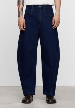 Person trägt dunkelblaue Jeans mit weitem Bein und braune Schuhe und steht mit entspannten Händen seitlich vor einem einfarbigen Hintergrund.