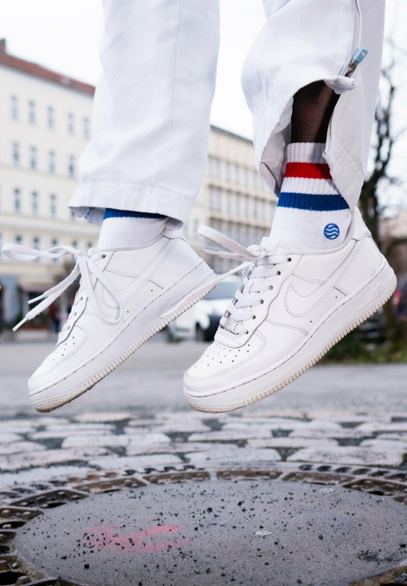Valged Nike Air Force 1 tossud perforatsioonidega varvaste osas ja nööpidega. Kandmine valgete, siniste ja punaste triibuliste sokkidega. Hüppamine hobukivist tänaval.