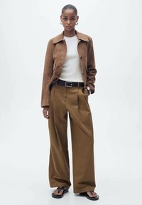 Braune Wildlederjacke, cremefarbener Pullover und hochgeschnittene braune weite Hosen. Ein schwarzer Gürtel und flache Sandalen vervollständigen das Outfit.