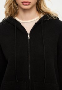 Černá mikina na zip s texturovanou tkaninou, šňůrkami a stříbrným zipem. Přes lehký béžový top, který ukazuje čistý výstřih.