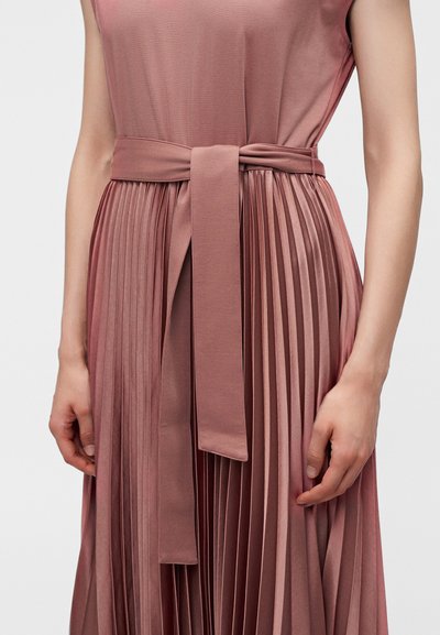 Robe sans manches rose poudré avec une jupe plissée et une large ceinture en tissu nouée à la taille, portée par une personne debout.