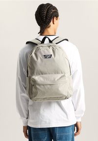 Mochila gris hecha de tela, con un bolsillo frontal, correas acolchadas para los hombros y un parche con el logo en la parte superior. Diseño sencillo sin patrones.