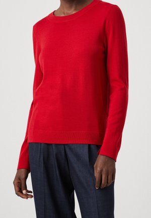 Person trägt einen schlichten roten Pullover mit langen Ärmeln und dunkelblaue, maßgeschneiderte Hosen, gezeigt von Hals bis Hüfte vor einem einfarbigen Hintergrund.