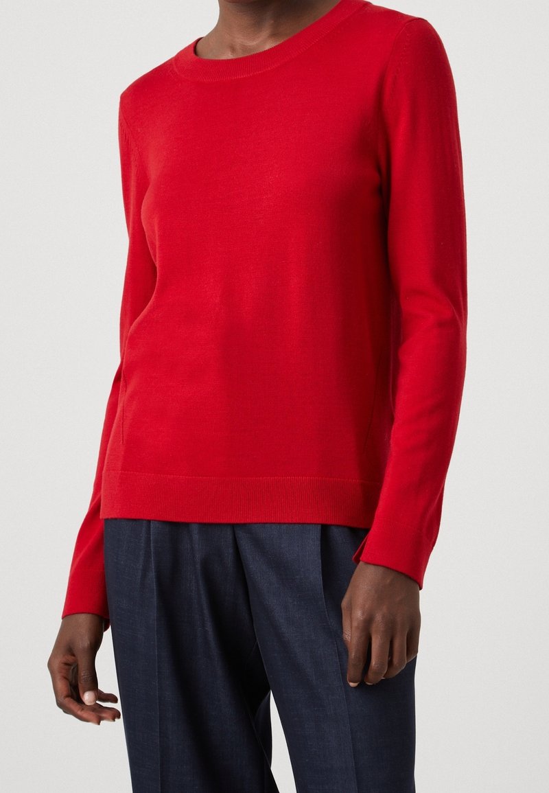 Personne portant un pull rouge uni à manches longues associé à un pantalon tailleur bleu marine foncé, montré du cou aux hanches sur un fond uni.