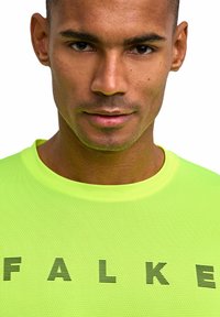 Fluorescencyjna żółta koszulka z krótkim rękawem o siateczkowej fakturze. Na przodzie duży napis „FALKE” w czarnych, prążkowanych literach. Widok z bliska.
