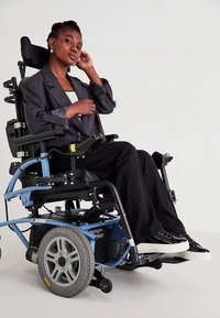 Cadeira de rodas elétrica com estrutura azul, assento preto e rodas traseiras grandes. O utilizador veste um blusão cinza e calças pretas, sentado confortavelmente.