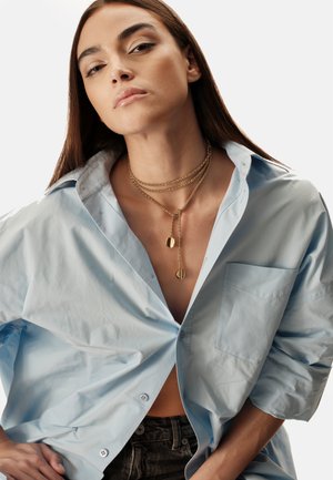 Camisa de botones de gran tamaño en azul claro con un bolsillo único, combinada con collares de cadenas doradas en capas que llevan colgantes redondos.