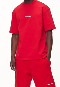 Punainen puuvillainen t-paita ja siihen sopivat shortsit, joissa on pyöreä kaula-aukko ja vasemman rinnan yläpuolella valkoinen "PEGADOR"-logo.