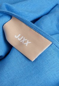 Tela azul clara con una textura suave, que presenta una etiqueta beige con "JJXX" en letras blancas. La tela está bien doblada.