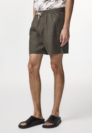 Olivgrüne Leinenshorts mit elastischem Bund und Kordelzug. Kombiniert mit schwarzen Slippern, die ein glattes, offenes Design haben.