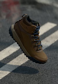 Brązowy but trekkingowy z wysokim kołnierzem, z niebieskimi sznurówkami i metalowymi oczkami. Gumowa podeszwa zapewniająca przyczepność. Gładkie i teksturowane materiały górne.