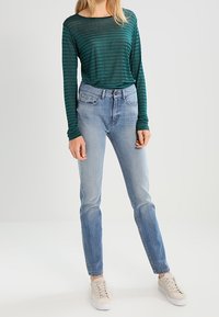Turanstrimmig långärmad topp i teal som kombineras med ljusblå jeans och beigefärgade sneakers. Slim fit jeans med lätt förstoring; toppen har en mjuk textur.