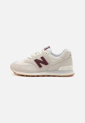 Hellbeiger Wildleder-Sneaker mit bordeauxrotem „N“-Logo, weißen Schnürsenkeln, gepolstertem Kragen und Gummisohle, seitlich dargestellt.