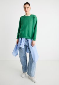 Sweat-shirt vert à col rond, superposé à une chemise rayée bleue et blanche. Jeans amples bleu clair et baskets blanches complètent la tenue.