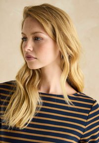 Femme aux longs cheveux blonds portant un haut rayé bleu marine et marron, regardant vers la gauche sur un fond neutre.