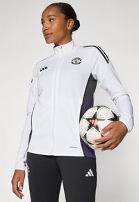 Veste de sport blanche avec des panneaux latéraux gris, dotée de accents noirs et d'un logo. Pantalons noirs avec le logo Adidas blanc. Le joueur tient un ballon de football à motifs.