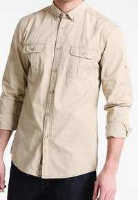 Camicia beige con colletto, due tasche sul petto, maniche arrotolate e bottoni marroni. Il tessuto è liscio con una leggera texture.