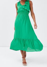 Robe verte sans manches avec un décolleté à volants, une taille smockée et un ourlet à étages. Tissu léger, design fluide et longueur au niveau des genoux.