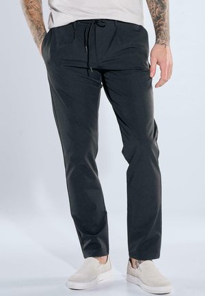 Man wearing black drawstring pants and white slip-on shoes, standing against a plain light background with hands relaxed by sides.