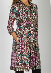 Kobieta w długiej sukience z kolorowym geometrycznym wzorem, stojąca z jedną ręką w kieszeni na jednolitym tle.