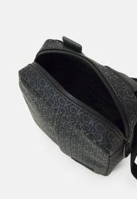 Borsa a tracolla nera con motivo a logo in rilievo. Presenta un'apertura con zip, una superficie liscia e una tracolla regolabile. L'interno è foderato in nero.