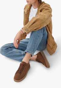 Hnědé semišové tenisky s kulatou špičkou, texturovaným svrškem a šněrováním. Umístěné na světlých džínách, s viditelnými detaily z béžového semiše na bundě.