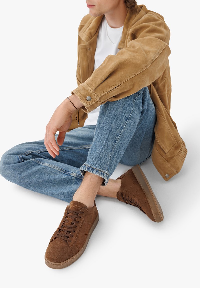 Hnědé semišové tenisky s kulatou špičkou, texturovaným svrškem a šněrováním. Umístěné na světlých džínách, s viditelnými detaily z béžového semiše na bundě.