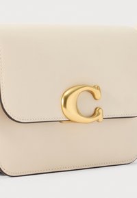 Borsa in pelle beige con una chiusura dorata che presenta una "C" stilizzata. Texture liscia, forma rettangolare e dettaglio di cucitura a contrasto.