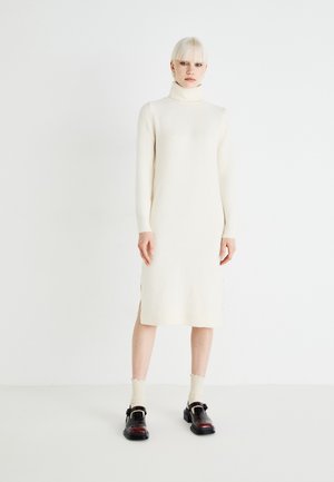 Robe pull crème avec un col haut, des manches longues, des fentes latérales et une texture douce. Associée à des chaussures noires et des chaussettes de couleur claire.