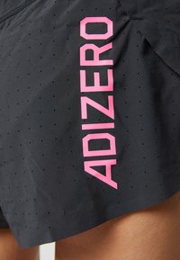 Shorts en tissu noir perforé avec un texte vertical "ADIZERO" rose vif sur le côté, portés par une personne avec la peau de la cuisse visible.