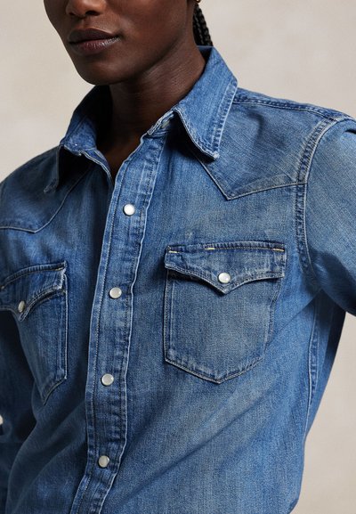 Prim-plan al unei persoane care poartă o cămașă din denim albastru cu nasturi tip capse și două buzunare la piept cu clapete.