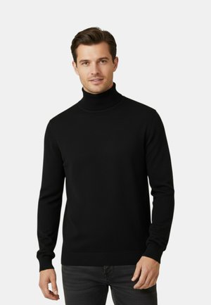 Uomo con maglione nero a collo alto e jeans scuri in piedi davanti a uno sfondo chiaro e uniforme, rivolto verso l'obiettivo con un leggero sorriso.