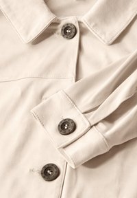 Beige fake mockasin jacka med spetsig krage, knappstängningar och uppvikkta ärmslut. Har metallknappar med ingraverade textdetaljer.