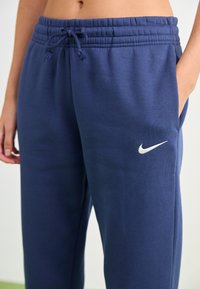 Nærbilde av en person iført marineblå Nike joggebukser med snor i livet og hvit Swoosh-logo på venstre lår, hånden hvilende i lomma.