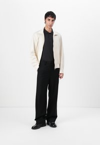 Ljusbeige zip-jacka med en slät textur, tillsammans med en svart polo skjorta och breda svarta randiga byxor. Svarta skor fulländar looken.