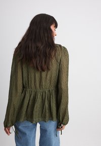 Blouse verte olive, texturée, à manches longues, avec une taille froncée et des liens aux poignets. Design fluide, tissu semi-transparent et coupe décontractée.