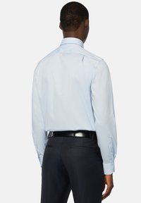 Chemise bleu clair à manches longues, coupe ajustée, avec poignets à boutons, associée à un pantalon sombre et une ceinture noire, vue de l'arrière.
